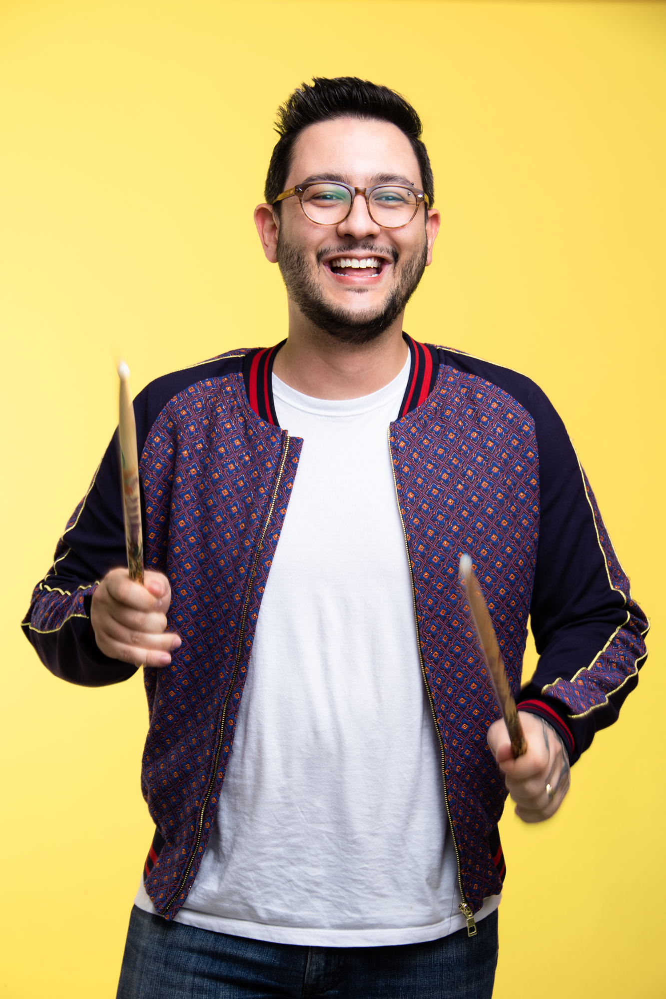 A picture of me, Paulo Cholla, wearing a purple jacket with some details, and a white t-shirt, smiling and holding drumsticks, against a yellow background
